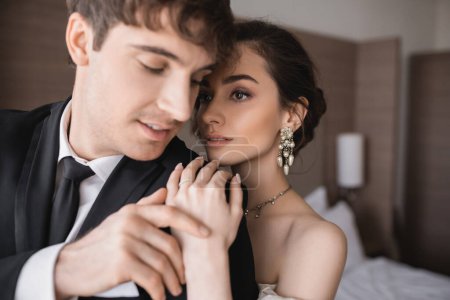 stunning and young bride with brunette hair, in elegant jewelry hugging shoulder of groom in classic formal wear with tie while standing together in modern hotel room after wedding ceremony 