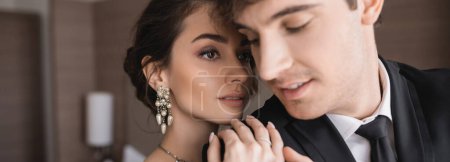 pretty and young bride with brunette hair, in elegant jewelry hugging shoulder of groom in classic formal wear with tie while standing together in modern hotel room after wedding ceremony, banner