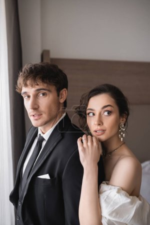 bride with brunette hair, in elegant jewelry looking away and standing in white dress and hugging shoulder of groom in classic formal wear with tie in modern hotel room after wedding ceremony 