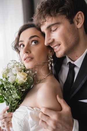 glücklicher Bräutigam in klassisch-formaler Tracht rührende Hand der hübschen jungen Braut in Schmuck, weißes Kleid mit Brautstrauß beim Zusammenstehen im modernen Hotelzimmer nach der Trauung 