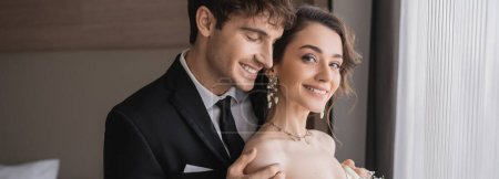 happy groom in classic formal wear touching shoulder of elegant and cheerful young bride in jewelry with pearls while standing together in modern hotel room after wedding ceremony, banner 