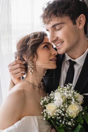 fröhlicher Bräutigam in klassischer formeller Kleidung umarmt glückliche Braut in Schmuck, weißes Kleid mit Brautstrauß beim Zusammenstehen im modernen Hotelzimmer nach der Trauung 