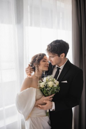 Bräutigam in klassischer formeller Kleidung umarmt glückliche Braut in Schmuck, weißes Kleid mit Brautstrauß, während sie nach der Trauung im modernen Hotelzimmer zusammensteht 
