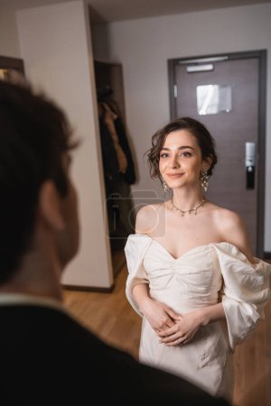 pretty young bride in elegant jewelry and white dress looking at blurred groom while standing near entrance door to modern hotel room after wedding ceremony, happy couple 