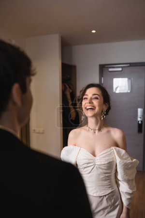 hübsche junge Braut in elegantem Schmuck und weißem Kleid, die lachend den verschwommenen Bräutigam ansieht, während sie nach der Trauung in der Nähe der Eingangstür zum modernen Hotelzimmer steht, glückliches Paar 