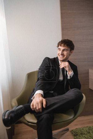 homme heureux en noir vêtements formels avec cravate et chaussures classiques assis sur un fauteuil confortable et regardant loin le jour du mariage, reposant dans la chambre d'hôtel moderne, beau marié 
