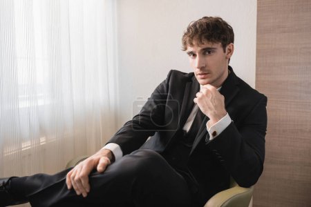 Foto de Hombre de éxito en ropa formal negro con corbata sentado en un sillón cómodo y mirando a la cámara en el día de la boda, descansando en la habitación de hotel moderna, novio guapo - Imagen libre de derechos