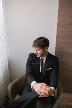 homme heureux en costume noir avec cravate souriant et assis avec les mains serrées sur fauteuil confortable et regardant loin le jour du mariage, se reposant dans la chambre d'hôtel moderne, beau marié 