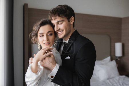 happy groom in classic formal wear touching hand of elegant young bride in jewelry and white dress while standing together in modern hotel room during their honeymoon after wedding 