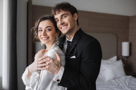 glücklicher Bräutigam in klassisch-formaler Kleidung umarmt elegante junge Braut in Schmuck und weißem Kleid, während sie während ihrer Flitterwochen nach der Hochzeit gemeinsam in die Kamera im modernen Hotelzimmer schauen 
