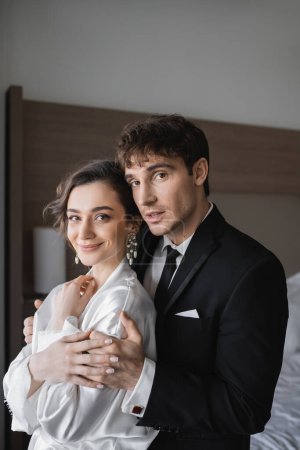 Bräutigam in klassischer formeller Kleidung umarmt glückliche junge Braut in Schmuck und weißem Kleid, während sie zusammen in einem modernen Hotelzimmer während ihrer Flitterwochen nach der Hochzeit stehen 