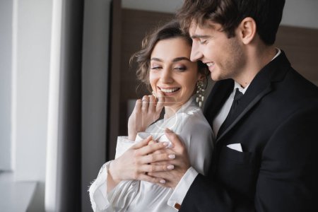 Bräutigam im klassischen schwarzen Anzug umarmt glückliche junge Braut in Schmuck und weißem Kleid, während sie zusammen in einem modernen Hotelzimmer während ihrer Hochzeitsreise am Hochzeitstag steht, freudiges Brautpaar 