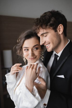 Bräutigam mit geschlossenen Augen im klassischen schwarzen Anzug umarmt glückliche junge Braut in Schmuck und weißem Brautkleid, während sie zusammen in einem modernen Hotelzimmer während ihrer Flitterwochen stehen, Brautpaar 
