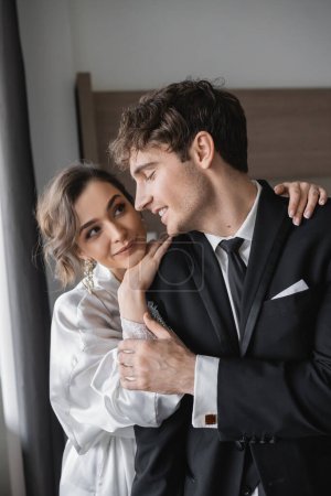 Photo for Pleased bride in jewelry and white silk robe leaning on shoulder of joyful groom in classic black suit while standing together in modern hotel room during honeymoon, newlyweds - Royalty Free Image