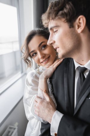 happy bride in jewelry and white silk robe leaning on shoulder of blurred groom in classic black suit while standing together in modern hotel room during honeymoon, newlyweds  Poster 654954938