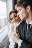 happy bride in jewelry and white silk robe leaning on shoulder of blurred groom in classic black suit while standing together in modern hotel room during honeymoon, newlyweds  Sweatshirt #654954938