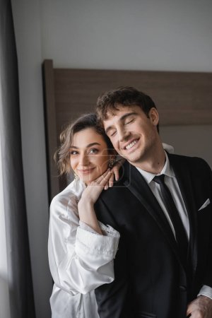 happy bride in jewelry and white silk robe leaning on shoulder of joyful groom in classic black suit while standing together in modern hotel room during honeymoon, newlyweds 