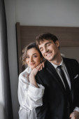 happy bride in jewelry and white silk robe leaning on shoulder of joyful groom in classic black suit while standing together in modern hotel room during honeymoon, newlyweds  Poster #654954944