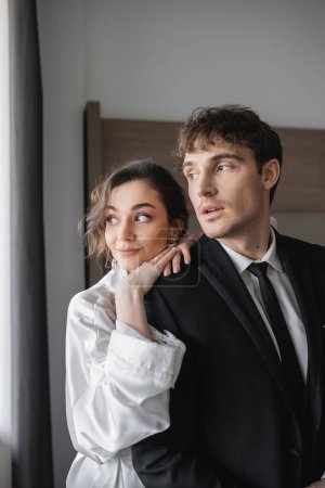 young bride in jewelry and white silk robe leaning on shoulder of groom in classic formal wear while standing and looking away together in modern hotel room during honeymoon, newlyweds 