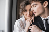 charming bride in jewelry and white silk robe leaning on shoulder of groom in classic formal wear while standing together in modern hotel room during honeymoon, newlyweds  puzzle #654954980