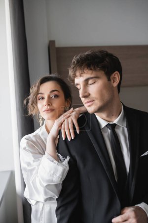 pretty bride in jewelry and white silk robe leaning on shoulder of groom with closed eyes in classic formal wear while standing together in modern hotel room during honeymoon, newlyweds 