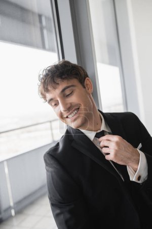 happy man in classic formal wear adjusting black tie while doing preparations and standing in modern hotel room near window, groom on wedding day, special occasion  