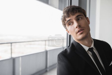 homme rêveur en tenue classique avec cravate noire et chemise blanche debout dans la chambre d'hôtel moderne et regardant la fenêtre, marié le jour du mariage, occasion spéciale  