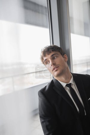 thoughtful man in classic formal wear with black tie and white shirt standing in modern hotel room and looking at window, groom on wedding day, special occasion  