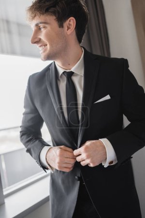 Foto de Hombre feliz en ropa formal clásica con corbata negra y camisa blanca abotonando chaqueta y de pie en la habitación de hotel moderna cerca de la ventana, novio en el día de la boda, ocasión especial - Imagen libre de derechos