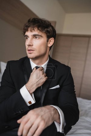handsome man in classy formal wear with white shirt adjusting black tie while looking away in modern hotel room, groom on wedding day, special occasion  