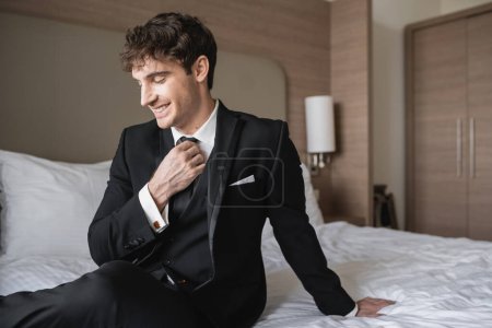 cheerful man in classy formal wear with white shirt adjusting black tie while looking away and sitting on bed in modern hotel room, groom on wedding day, special occasion  