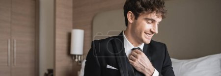 Photo for Cheerful man in classy formal wear with white shirt adjusting black tie while looking away in modern hotel room, groom on wedding day, special occasion, banner - Royalty Free Image