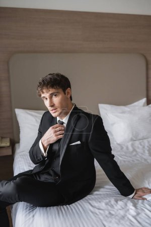 good looking man in classy formal wear with white shirt adjusting black tie while looking at camera and sitting on bed in modern hotel room, groom on wedding day, special occasion  