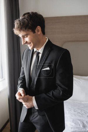 Porträt eines glücklichen jungen Mannes im klassischen schwarzen Anzug mit Krawatte und weißem Hemd, der im modernen Hotelzimmer lächelt, Bräutigam am Hochzeitstag, besonderer Anlass  