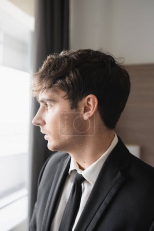 Photo for Portrait of good looking man in classy formal wear with black tie and white shirt looking at window in modern hotel room, groom on wedding day, special occasion, side view - Royalty Free Image