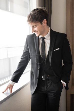 glücklicher Mann in eleganter formaler Kleidung mit schwarzer Krawatte und weißem Hemd, der mit der Hand in der Tasche auf der Fensterbank in einem modernen Hotelzimmer neben dem Fenster steht, Bräutigam am Hochzeitstag, besonderer Anlass 