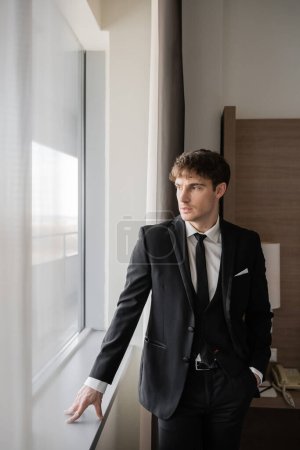 schöner Mann in eleganter formaler Kleidung mit schwarzer Krawatte und weißem Hemd, der mit der Hand in der Tasche auf der Fensterbank in einem modernen Hotelzimmer in Fensternähe steht, Bräutigam am Hochzeitstag, besonderer Anlass 