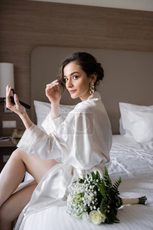 attraktive Frau in weißem Seidenmantel, die sich auf ihre Hochzeit vorbereitet, während sie Make-up macht und einen Taschenspiegel hält, auf dem Bett neben Brautstrauß im Hotelzimmer sitzt, besonderer Anlass, junge Braut