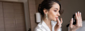 attractive bride in white silk robe preparing for her wedding while touching lips, holding pocket mirror in hotel room on wedding day, special occasion, banner  mug #654955278