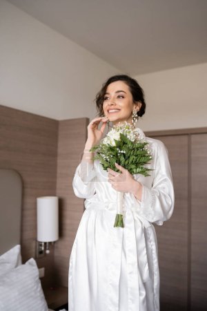 young woman with brunette hair in white silk robe and pearl earrings holding bridal bouquet while preparing for her wedding in hotel room, special occasion, happy bride 