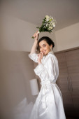 alluring young woman with brunette hair in white silk robe and pearl earrings holding bridal bouquet while preparing for her wedding in hotel room, special occasion, happy bride  t-shirt #654955310