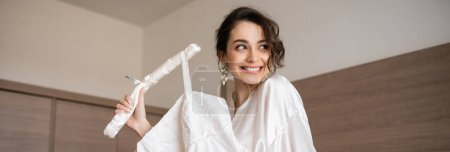 happy young bride with brunette hair in white silk robe holding soft hanger with elegant wedding dress and smiling in bedroom of hotel room, special occasion, charming woman, banner 