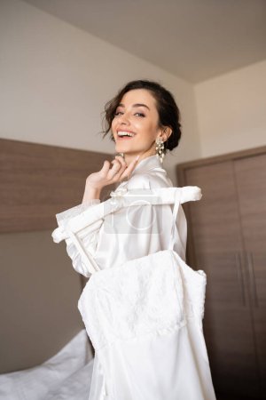 Foto de Novia alegre con pelo morena en túnica de seda blanca sosteniendo percha suave con elegante vestido de novia y sonriendo en el dormitorio de la habitación de hotel, ocasión especial, mujer hermosa - Imagen libre de derechos