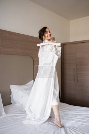 happy young bride with brunette hair in white silk robe holding soft hanger with elegant wedding dress and standing on bed in bedroom of hotel room, special occasion, pretty woman 