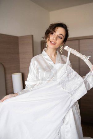 joven novia con pelo morena en túnica de seda blanca sosteniendo percha suave con elegante vestido de novia en el dormitorio de la habitación de hotel, ocasión especial, mujer encantadora 