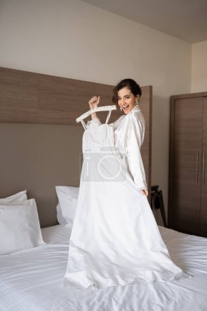 Foto de Novia excitada con cabello moreno de pie en bata de seda blanca sosteniendo percha suave con elegante vestido de novia y de pie en la cama en el dormitorio de la habitación de hotel, ocasión especial, mujer encantadora - Imagen libre de derechos