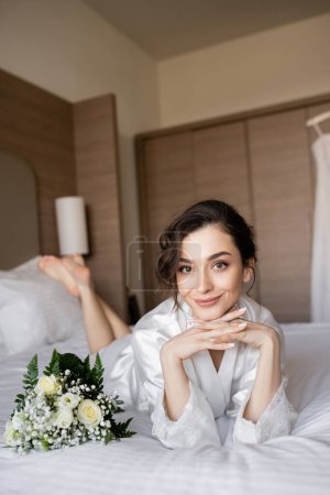 charming woman with brunette hair lying in white silk robe with clenched hands next to bridal bouquet on bed in hotel room on wedding day, special occasion, young bride