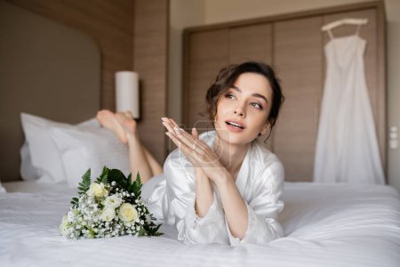 gorgeous woman with brunette hair and engagement ring on finger looking away and lying in white silk robe next to bridal bouquet on bed with wedding dress on blurred background, young bride