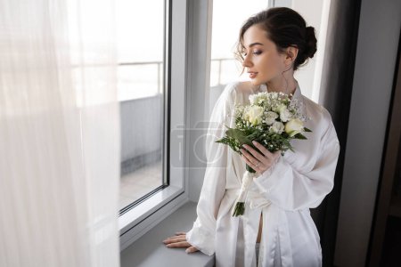 young woman with engagement ring on finger standing in white silk robe and holding bridal bouquet next to tulle curtain and window in hotel suite, special occasion, bride on wedding day