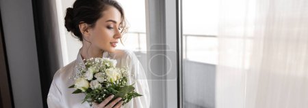 Photo for Young woman with brunette hair standing in white silk robe and holding bridal bouquet next to tulle curtain and window in hotel suite, special occasion, bride on wedding day, banner - Royalty Free Image